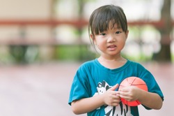 Happy cute asian girl smiling and  holding small basketball in the park with sun light background. Funny adorable Asia child playing mini ball alone in the garden with blurred background.