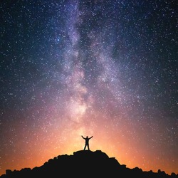 Man and the Universe. A person is standing on the top of the hill next to the Milky Way galaxy with his hands raised to the air. 