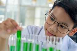 Selective focus, Asian scientist boy wearing eyeglasses smiling and looking green liquid in glass tubes in row to Analyzing record during science class at laboratory, copy space