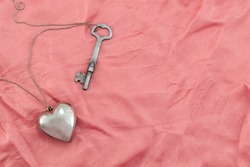 A silver heart and key on a chain, laying on rumpled pink fabric.
