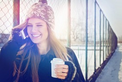 Closeup outdoor portrait of cute happy blonde Caucasian teenage girl with takeaway coffee talking on smartphone. Young woman with long hair with coat, gloves, knitted hat outdoors in winter by fence.