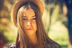 Beautiful young brunette woman with long hair flying in the wind and brown hat in park in summer. Head shot of gorgeous teenage girl with blue eyes. Retouched, filter, shallow depth of field.