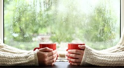 red circles in the hands of those dressed in sweaters against the background of a window with drops after the rain. a warming drink for a cozy conversation