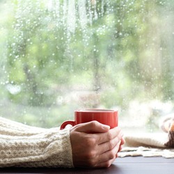 red mug with a hot drink in the hands of a person dressed in a warm sweater near the window / time to drink a warm drink