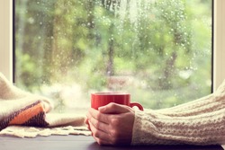 red mug of hot drink in hand, when behind is window is rain / cozy home atmosphere in autumn