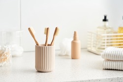 Cup with bamboo toothbrushes and stack of cotton towels placed on shelf near hygienic supplies in morning in bathroom