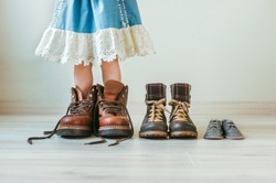 Close up of little girl putting on fathers hiking shoes. Three pairs of shoes for family indoor.