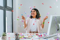 Young business woman having fun time catching confetti sitting at the desk in the office. Party time on the work place. Selective focus.