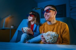 Father and daughter watching movie at home using 3D eye glasses and eating popcorn. Family activities. Selective focus on the male face. Blue light from the screen.