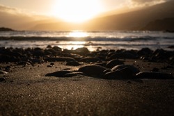 A mostly blurred closeup of black sand beach with volcanic rocks at sunset. Dramatic landscape. Golden hour at the sea shore. Warm evening by the ocean in Tenerife, the Canary Islands, Spain.
