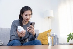 Sick Asian Woman Read Medicine Prescription on Her Mobilephone While Holding Pills Bottle on the Sofa