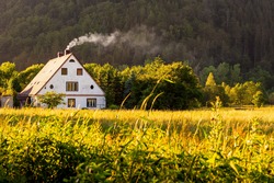 Rural view, a traditional polish country cottage house with smoke from the chimney next to pine forest. Rural cottage in the distance at sunset