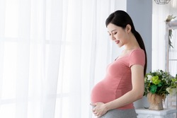 Side view of beautiful asian pregnant woman standing near window at home