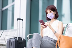 asian young businesswoman wearing face mask and sitting outside is using smart phone with suitcase on her business trip
