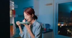 asian woman takes cake from opened refrigerator for late night supper in kitchen at night