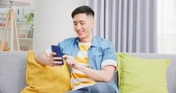 asian young man sitting on sofa check social media by mobile phone happily in living room at home