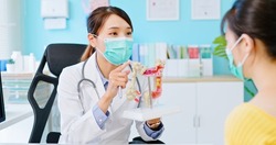 asian woman wears face mask to prevent COVID19 has colorectal cancer diagnosis in hospital - female doctor shows colonoscopy results and xray to patient on computer and explains by anatomical model