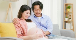 asian man is touching pregnant wife belly and expecting baby on couch at home