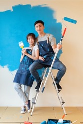 young asian couple decorate home - they painting wall with blue color together and smiling at you