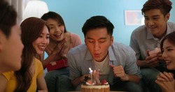 happy asian man surrounded by friends make wish with cake and blow out candles 