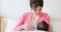 authentic shot of asian mother sitting on bed is breastfeeding her baby