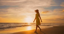 side view of asian woman wearing beautiful yellow dress walking by beach at golden sunset leaving footprints in sand - Female tourist on summer vacation