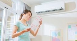 asian woman stand by air conditioner shocked and upset about electricity bill in hand at home - economic inflation concept
