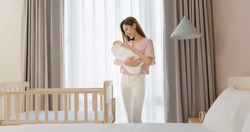 asian mother is hugging comforting cute newborn baby to sleep and standing besides crib at home