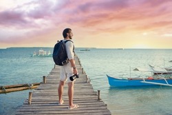 Photography and travel. Young man with rucksack holding camera standing on wooden fishing pier enjoying beautiful tropical sea view.