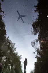 Blurred silhouette of reflection of one person walking alone on wet sidewalk of city park on rainy day. The plane flies across the sky. Abstract photography.