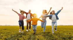 Happy young parents with daughters in casual clothes running with outstretched arms while enjoying time together on green meadow in summer evening