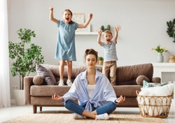 Happy mother with closed eyes meditating in lotus pose on floor trying to save inner harmony while excited children jumping on sofa and screaming in light spacious living room