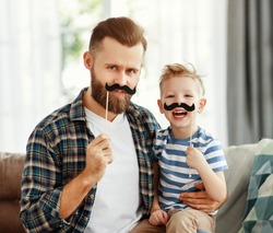 Happy young bearded man with little son having fun and playing with fake mustache while sitting together on sofa at home