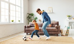 Full body bearded man in glasses trying to take ball from little boy in casual clothes while playing football near sofa at home together