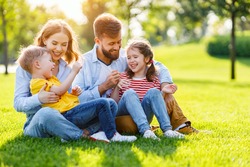 Cheerful parents smiling and playing with kids while sitting on green grass on sunny summer day in park