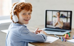 Happy boy in headphones smiling and looking at camera while making video call to teacher during online lesson at home