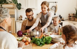 Positive young housewife with little daughters, husband and grandmother gathering around kitchen table and preparing delicious healthy dishes with fresh ingredients for family lunch at home