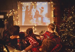 happy family in checkered pajamas: mother father and children watching projector, TV, movies with popcorn in christmas holiday evening   at home 