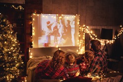 happy family in checkered pajamas: mother father and children watching projector, film, movies with popcorn in christmas holiday evening   at home 