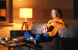 Happy woman in knitted sweater eating French fries and watching interesting movie while laughs and sitting cross legged on sofa in evening at home