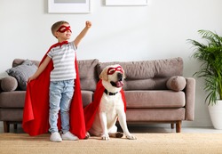 Full body of little boy in red superhero cloak and mask raising hand while playing with funny dog dressed in similar costume in living room at home