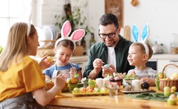 Joyful family wearing bunny ears headbands gathering at table in modern light kitchen and paining Easter eggs together