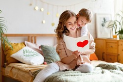 Cheerful mother hugging son and reading handmade greeting card with heart while resting on bed during holiday celebration mothers day  at home