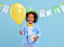Cheerful little black boy with curly hair in stylish suit and party hat smiling while standing against blue background decorated with colorful flags with yellow balloon and present in hands