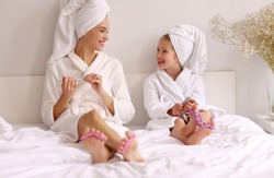Full body young mother and daughter in bathrobe and towels smiling and filing nails while resting on bed and doing manicure at home
