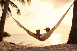 Full length of young woman, successful female freelancer using laptop while lying in hammock on the tropical beach at sunset, working remotely during summer vacation. Distance work concept