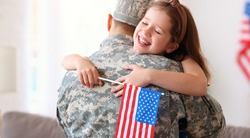 Happy little girl daughter with American flag hugging father in military uniform came back from US army, rear view of dad male soldier reunited  reunited with family at home