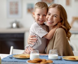 Optimistic woman embracing cheerful boy and looking at camera while sitting at table with cookies and milk and having breakfast in morning at home