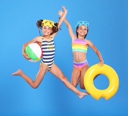 Two funny little girls sisters in swimsuits and swimming goggles jumping up in air with inflatable swimming ring and ball in hands on blue studio background, excited and ready for summer sea vacation
