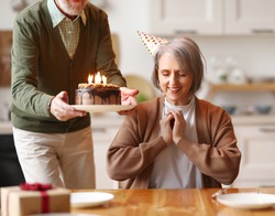 Happy elderly woman in party hat closing her eyes, she makes a wish, while her husband holds a chocolate cake with candles celebrating her Birthday at home, sitting together in kitchen